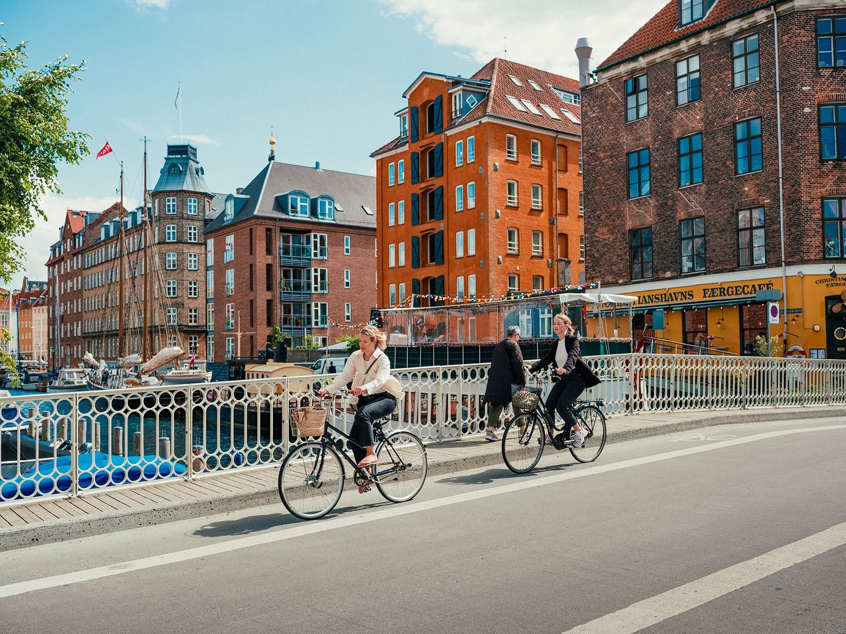 Zwei Frauen fahren mit dem Fahrrad über den Wilders Kanal; im Hintergrund viele große, rote Backsteingebäude