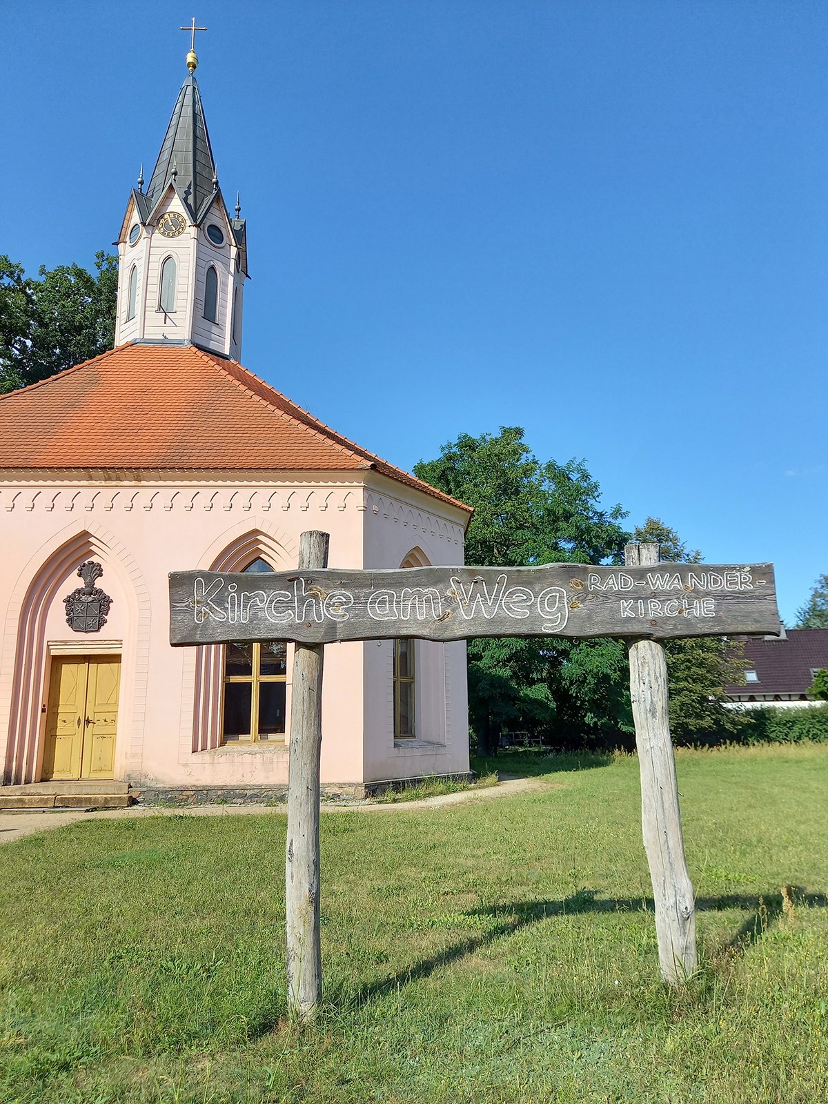 Holzschild mit der Aufschrift "Kirche am Weg" und im Hintergrund die Rad-Wander-Kirche